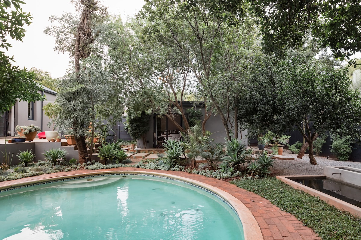 A serene pool area is surrounded by lush greenery and mature trees, creating a tranquil outdoor space. The pool's clear water reflects the surrounding foliage, while a path leads to the adjacent cottage, featuring soft landscaping and various plants.