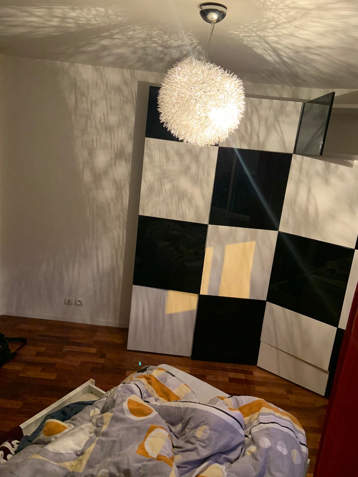 A bedroom space features a distinctive patterned wardrobe in black and white. Soft shadows are cast across the walls from a decorative light fixture above. A disheveled bed with a patterned duvet is present in the foreground, with wooden flooring visible throughout.