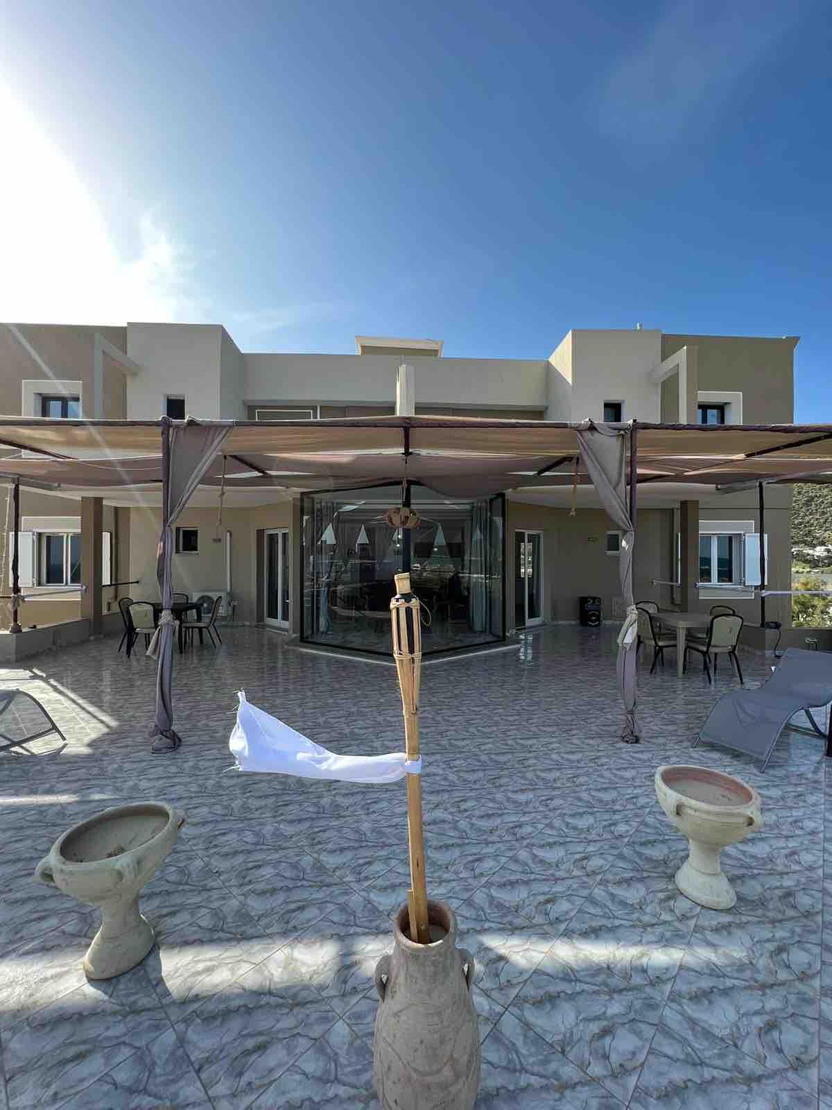 A spacious outdoor area features decorative stone planters and several lounge chairs under a canopy. The glass walls of the house create a seamless connection between the interior and exterior, while the bright sky adds a serene atmosphere to the setting.