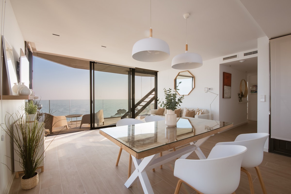 An open-plan living area features large glass walls that offer unobstructed views of the Mediterranean Sea. A glass-top dining table is positioned among modern white chairs, complemented by natural light and greenery from the plants. Cozy seating is visible in the background.