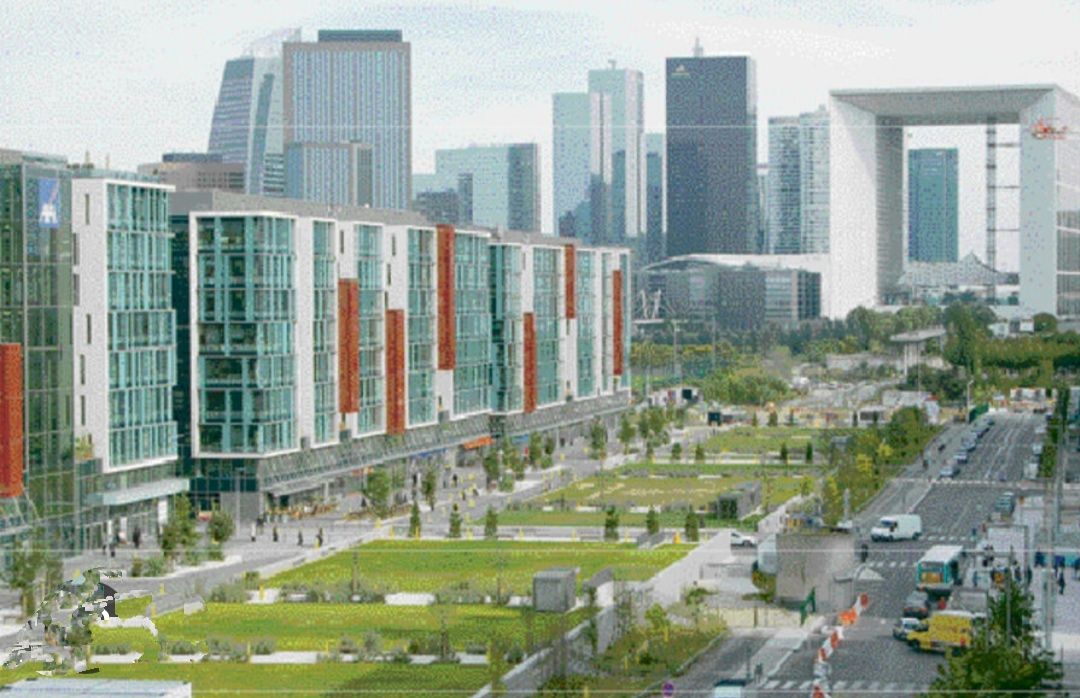 A vibrant urban scene is captured, featuring modern apartment buildings with colorful paneling alongside manicured green spaces. The backdrop showcases the iconic La Défense skyline with its distinctive arch, while pedestrians and vehicles populate the bustling streets.