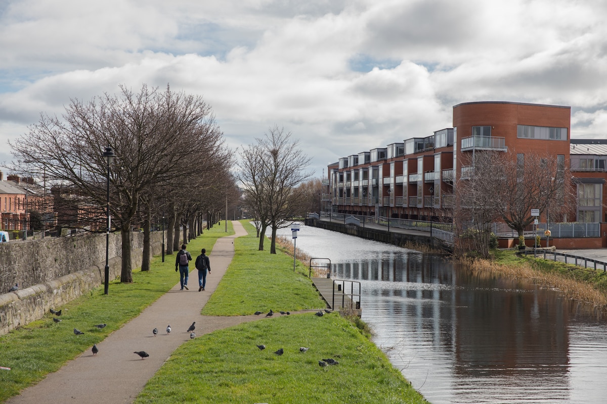 A scenic walkway is lined with trees alongside a tranquil waterway. Two individuals are seen strolling on a paved path, with lush green grass on either side. Modern residential buildings are visible in the background, reflecting the sky above and the water's surface.