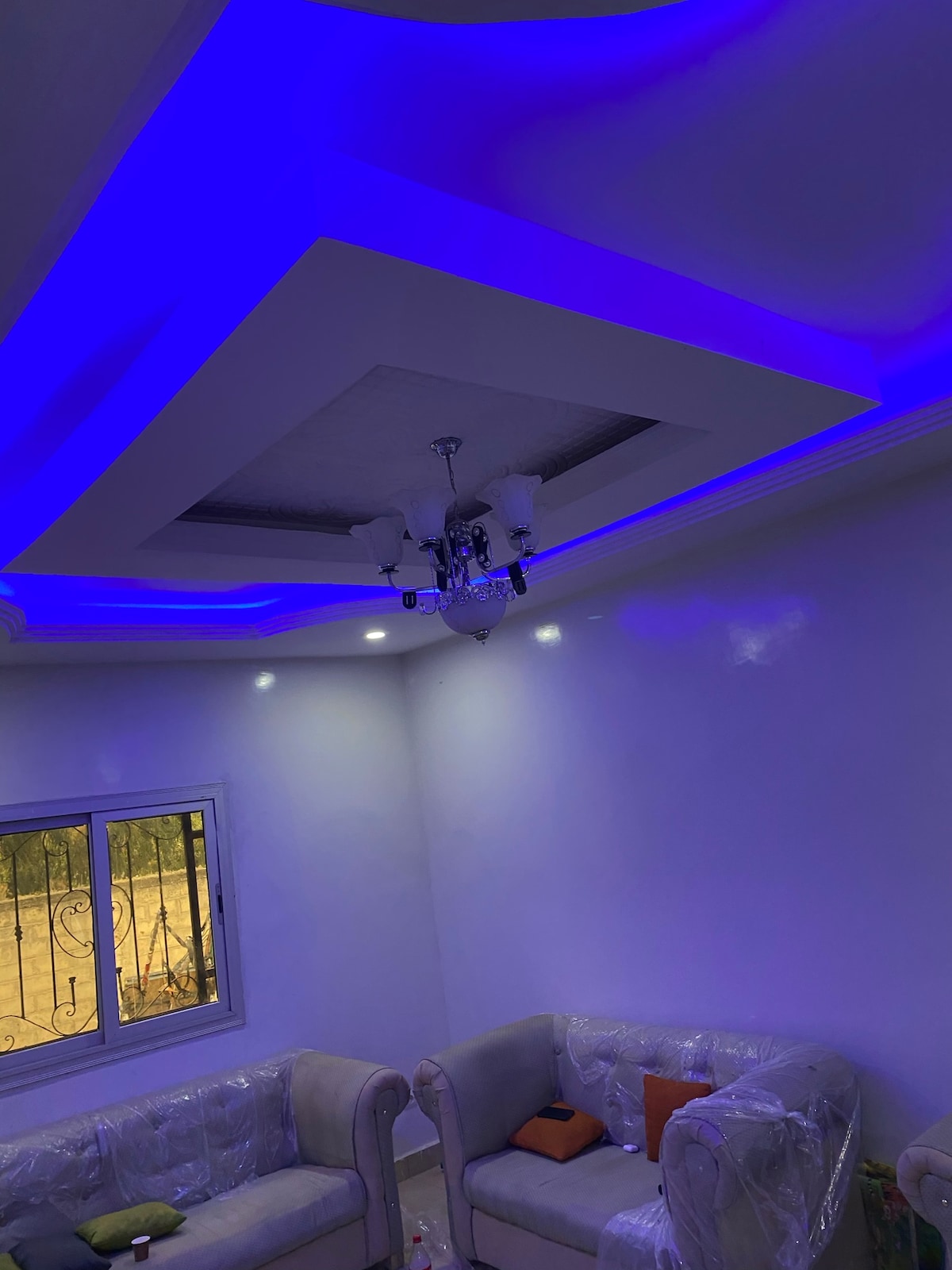 A softly illuminated ceiling is seen, featuring a modern chandelier with multiple lights. Blue ambient lighting enhances the room's corners. The window allows natural light in, revealing a view of greenery outside, while the beige seating adds comfort to the space.