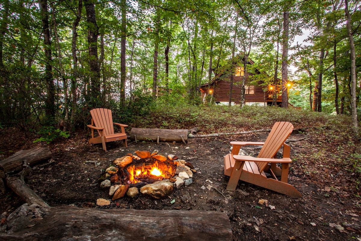 An outdoor fire pit is surrounded by two wooden adirondack chairs, offering a cozy seating area. The fire flickers within a circle of stones, while the cabin stands in the background, illuminated softly among the trees.