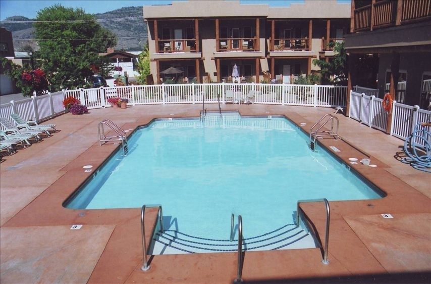 A clear, inviting swimming pool features steps leading into the water, surrounded by a spacious deck. Lounge chairs line the perimeter, alongside potted flowers. The backdrop includes a charming building with balconies. Lush greenery is visible in the vicinity, enhancing the serene environment.