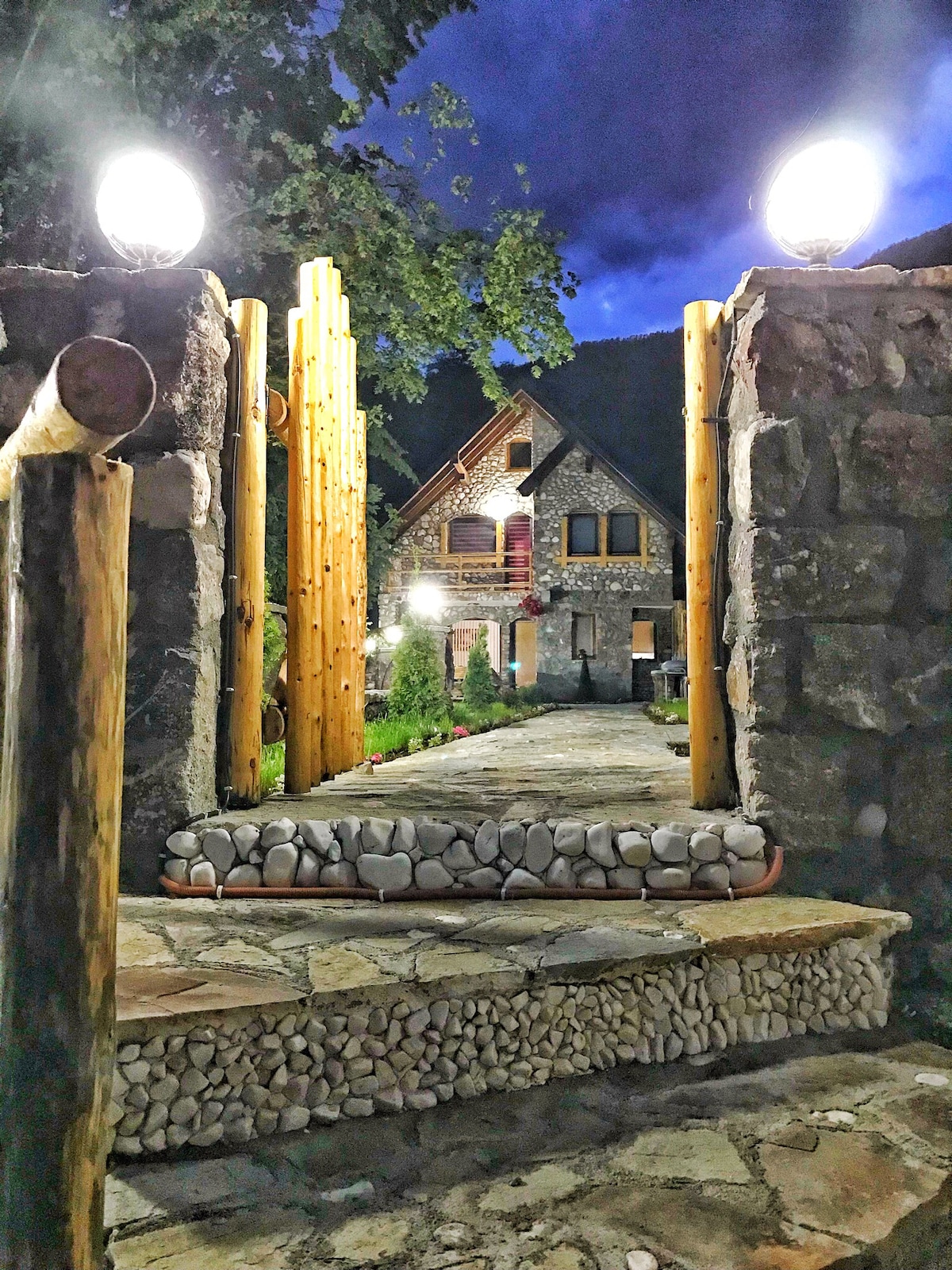 A stone pathway leads through wooden posts towards a chalet under a night sky. The entrance is framed by illuminated globes, and the surrounding greenery adds a natural touch to the scenic approach.