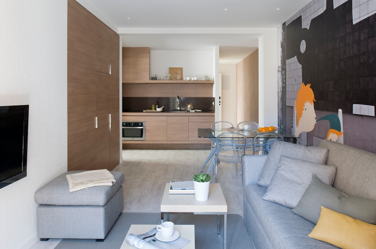 A modern living space is shown, featuring a soft gray sofa and a small white coffee table with a cup and orange. A dining area with transparent chairs is visible, along with a fully equipped kitchen in warm wood tones. A mural with colorful design adds character to the room.