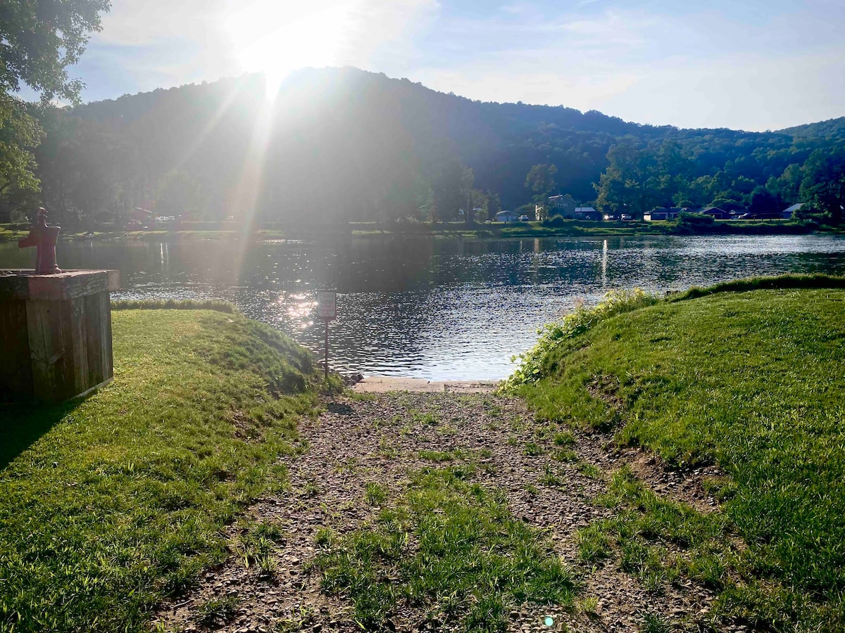 A serene view of the Allegheny River is captured, with sunlight reflecting off the water's surface. The foreground features a grassy slope leading down to the riverbank, bordered by pebbles. The surrounding landscape includes gently rolling hills, enhancing the tranquil atmosphere.