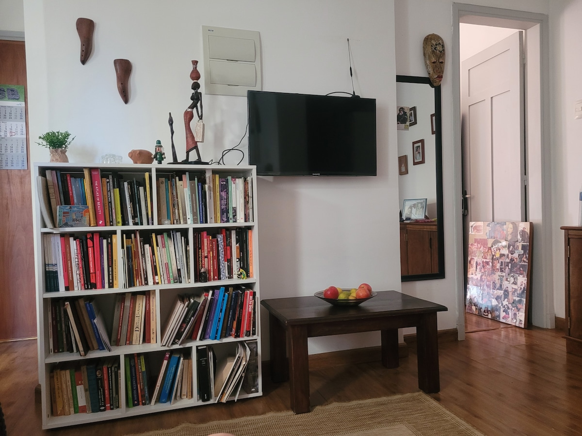 A neatly organized bookshelf filled with colorful books stands against the wall, complemented by a wooden coffee table with a bowl of fruit. A flat-screen television is mounted above the bookshelf, while a mirror and door are visible in the background.