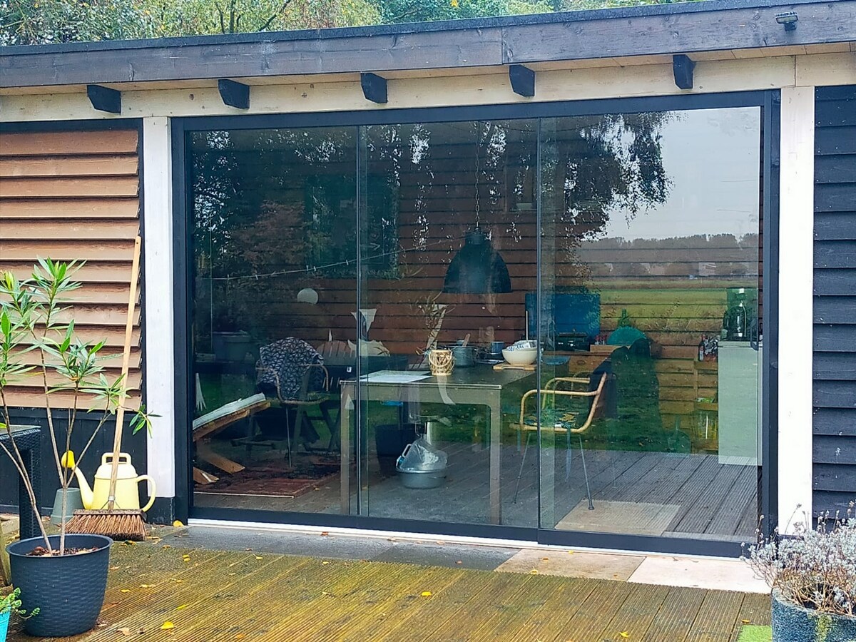 A modern facade with large glass sliding doors opens to a spacious living area featuring a dining table and chairs. Natural light fills the room, highlighting wooden accents and elegant furnishings. The outdoor deck and surrounding garden create an inviting connection to the lush environment beyond.