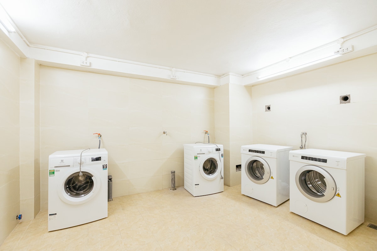 A designated laundry area features three white washing machines lined along a neutral-toned wall. The space is well-lit with overhead lighting, and a tiled floor extends throughout, offering a clean and functional environment for laundry needs.