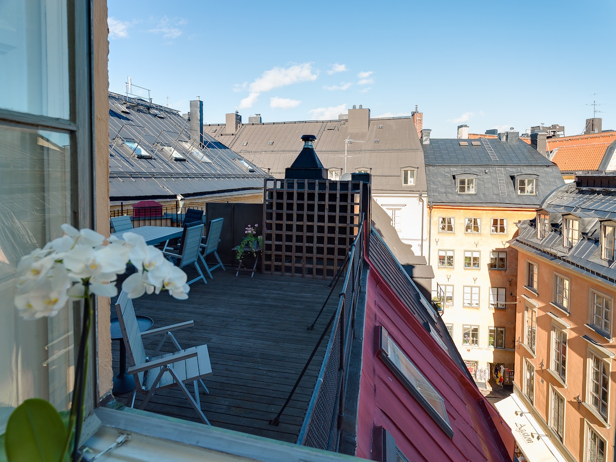 A private terrace is visible, surrounded by charming rooftops of Gamla Stan. Wooden decking is complemented by simple outdoor furniture, offering a space for relaxation. Bright skies with scattered clouds enhance the open atmosphere, while neighboring buildings display varied architectural styles.