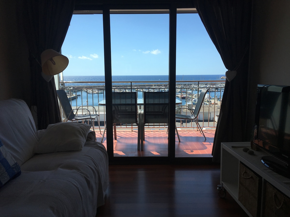 A wide view is presented through expansive glass doors, showcasing a sunny terrace overlooking the marina. Two outdoor chairs are positioned on the balcony, while a cozy white sofa is visible in the living area. Natural light fills the space, highlighting the serene coastal surroundings.