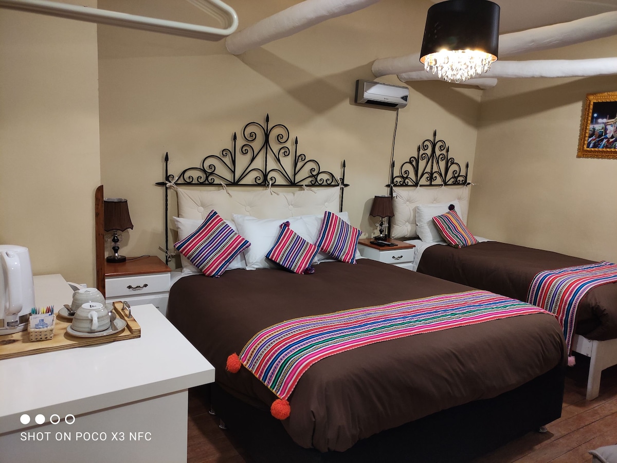 A comfortable sleeping area features two spacious beds with decorative headboards and colorful accent pillows. Nightstands with lamps are positioned beside each bed. A small tray holds tea-making supplies, providing convenient access to refreshments.