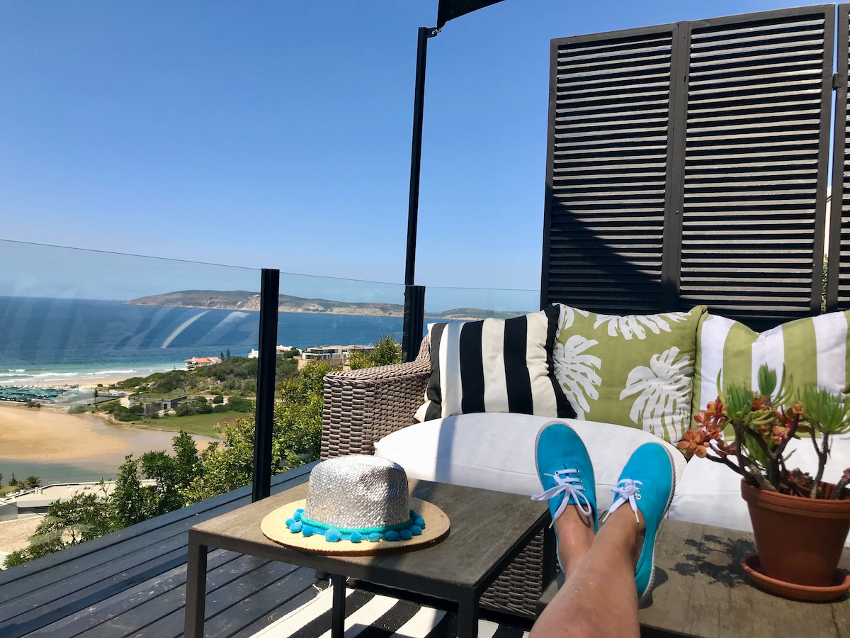 A comfortable seating area is featured on an outdoor terrace, showcasing a view of the bay. A patterned cushion lies atop grey seating, accompanied by a potted plant and a wide-brimmed hat on a round table. Bright blue skies frame the horizon.