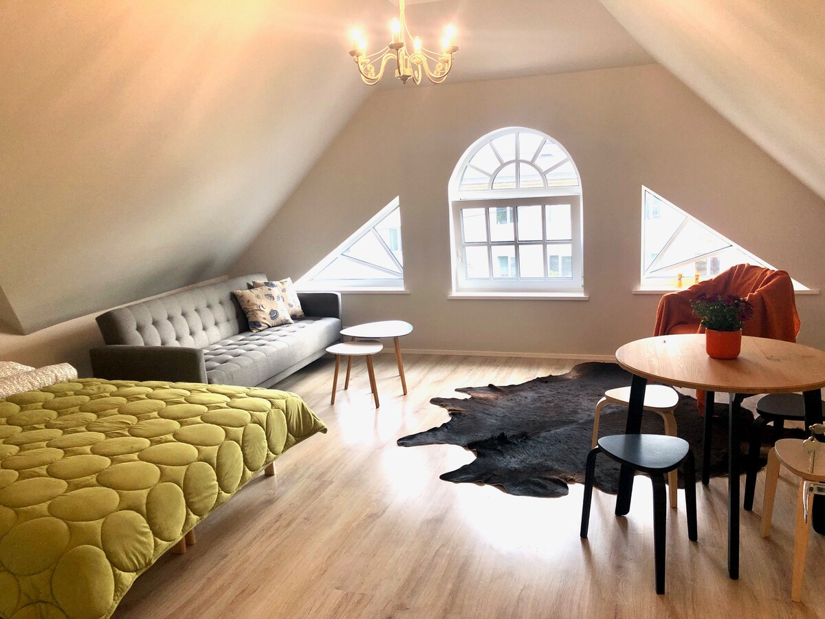 A cozy attic space features a comfortable sofa and a round wooden table surrounded by black chairs. A textured green bedspread is placed on a bed, while a potted plant adds a touch of nature. Large windows provide ample natural light.