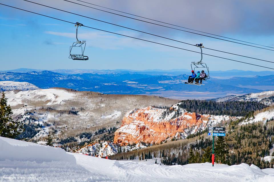 A scenic view of a ski area features chairlifts with passengers ascending the mountain. Snow-covered slopes stretch out below, bordered by pine trees and red rock formations. The expansive landscape reveals distant mountain ranges under a partly cloudy sky, creating a serene winter atmosphere.