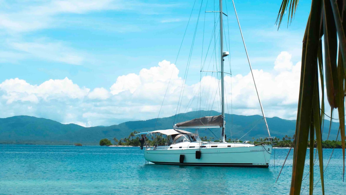 A modern sail yacht is anchored in calm turquoise waters, framed by lush green hills in the background. The bright blue sky above is dotted with soft white clouds, creating a serene atmosphere nearby the beautiful landscape.