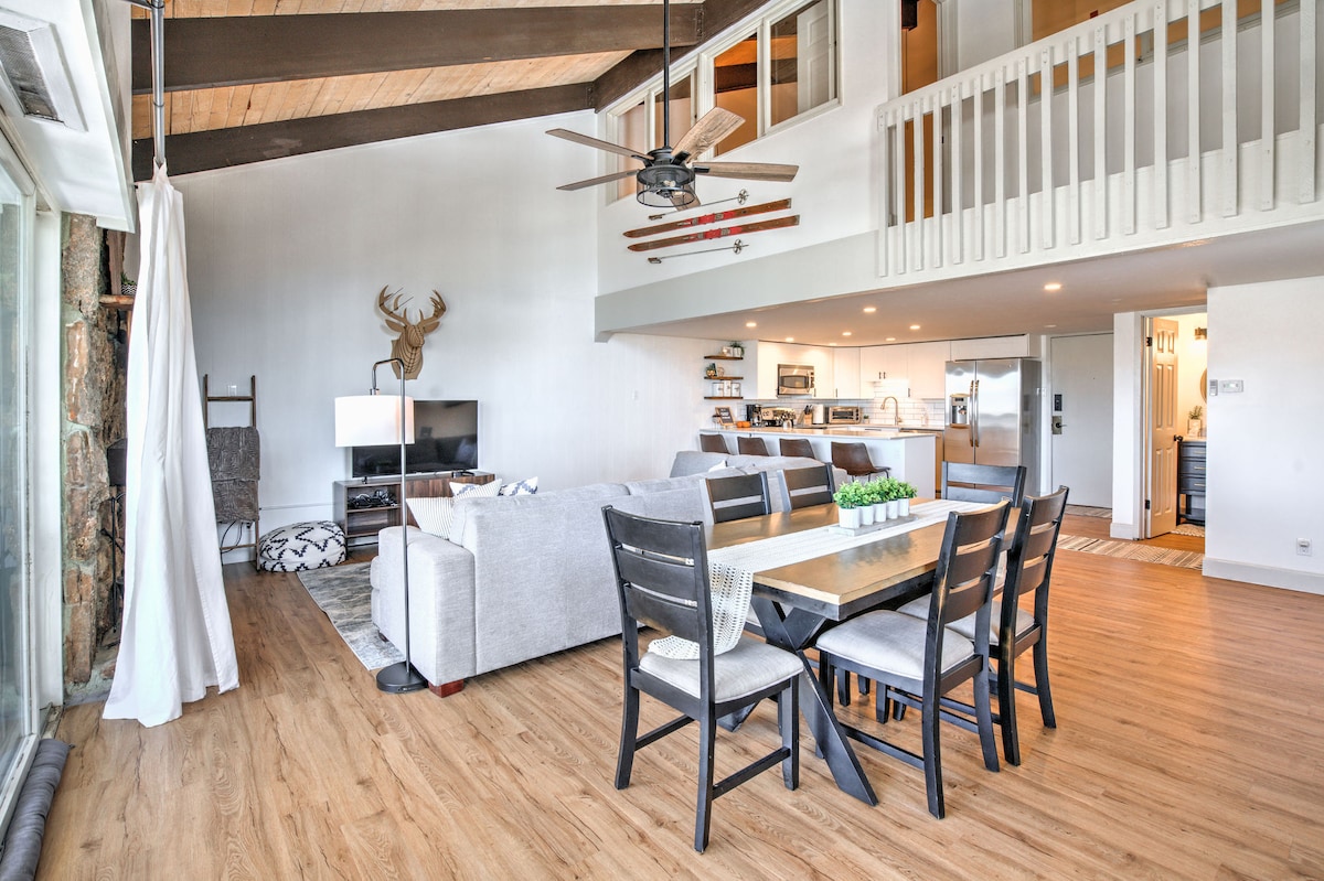 An open living area features a large sectional sofa facing a television on a sleek stand. A dining table with black chairs is positioned next to the well-equipped kitchen, creating a unified space. Exposed beams add character to the room, with natural light flooding in from large windows.