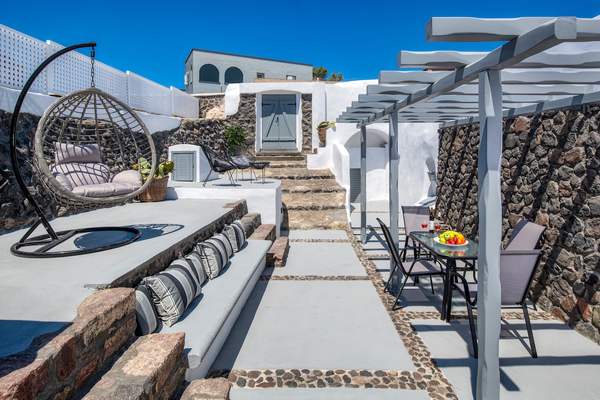 An outdoor area features a stonewall backdrop with a staircase leading to a door. A circular hanging chair and a dining set with gray chairs are present. Cushions are arranged on a raised platform, while a tray with fruits is placed on the table.