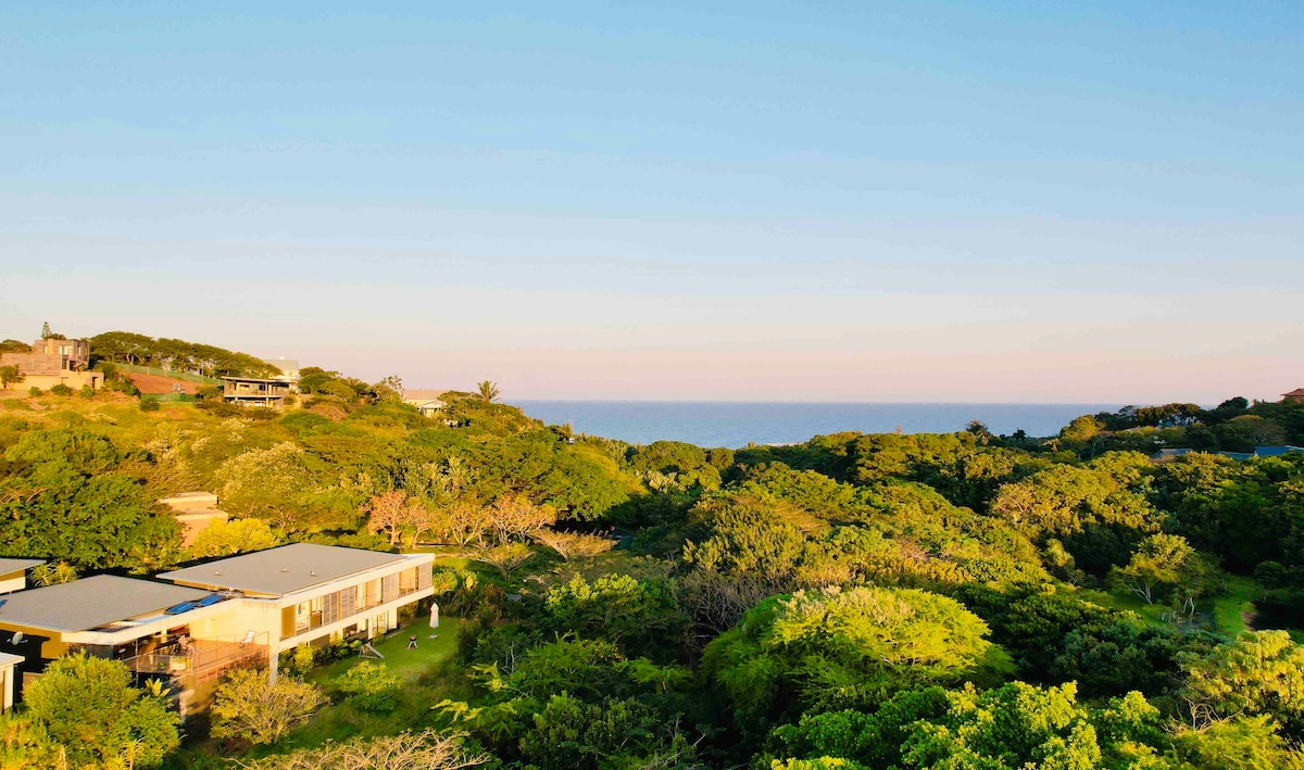 A panoramic view of a lush green landscape transitions to the ocean in the distance. The vegetation surrounds a modern two-story house with a flat roof, which is nestled harmoniously among the trees, creating a serene and natural setting.