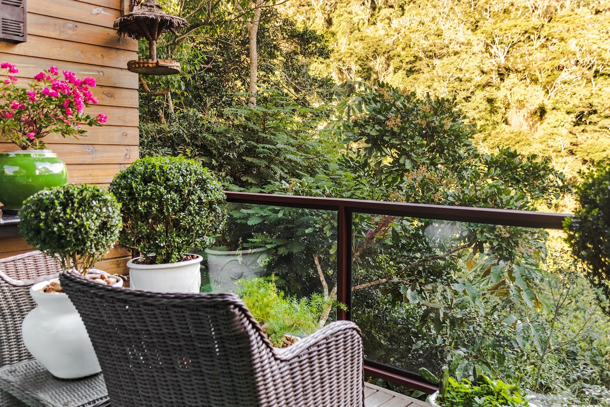 A balcony with two wicker chairs is positioned against a backdrop of lush greenery. Potted plants and vibrant flowers add color, while the surrounding trees create a serene atmosphere. The glass railing offers unobstructed views of the natural landscape.