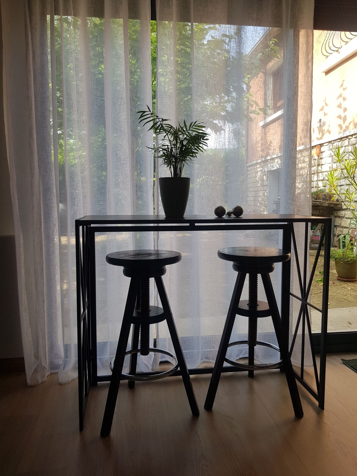 A narrow, modern bar table with two stools is positioned near sheer white curtains. A small potted plant adds a natural touch, while light filters through the curtains, illuminating the space and providing a glimpse of the outdoor area.