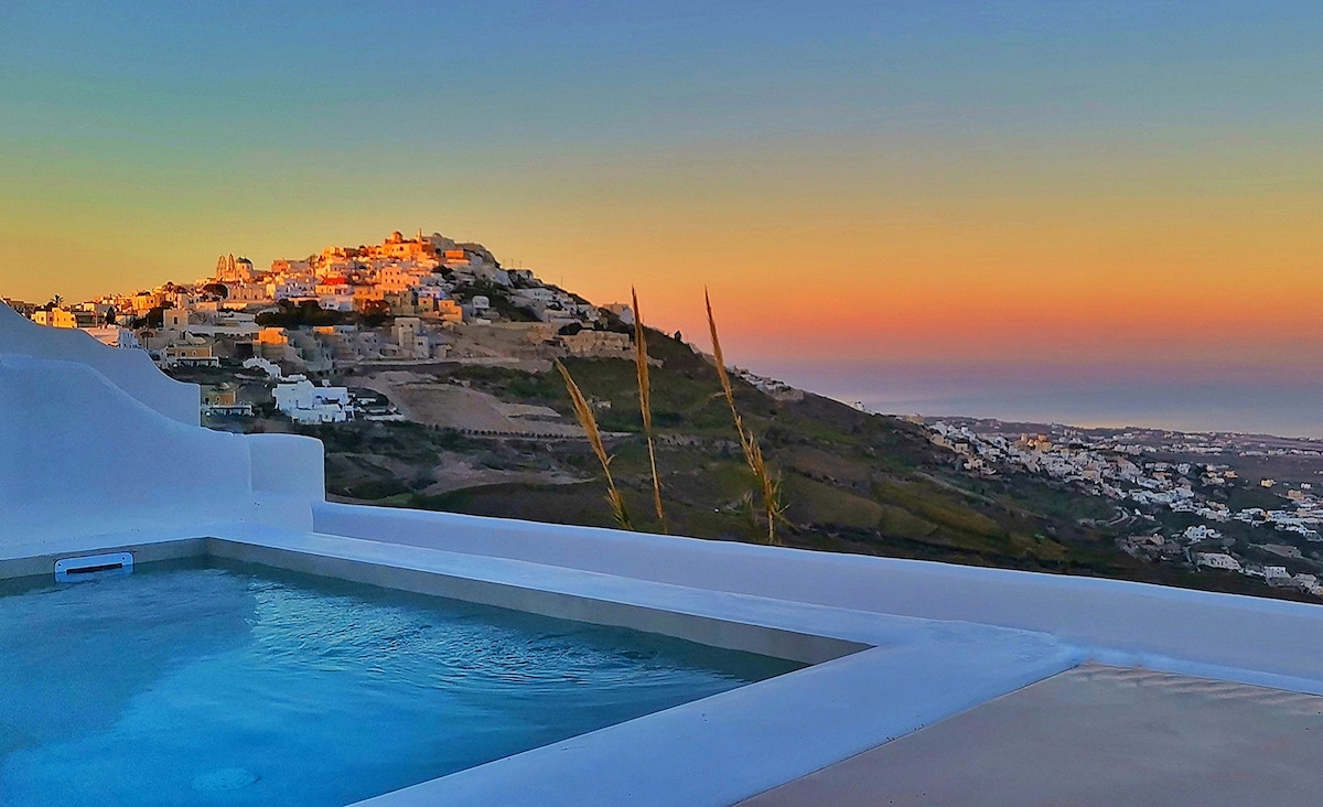 A heated mini pool is visible in the foreground, reflecting the soft hues of sunrise. The hillside village of Pyrgos can be seen in the distance, with its buildings illuminated by the early light. A gradient sky shifts from deep blue to warm orange.