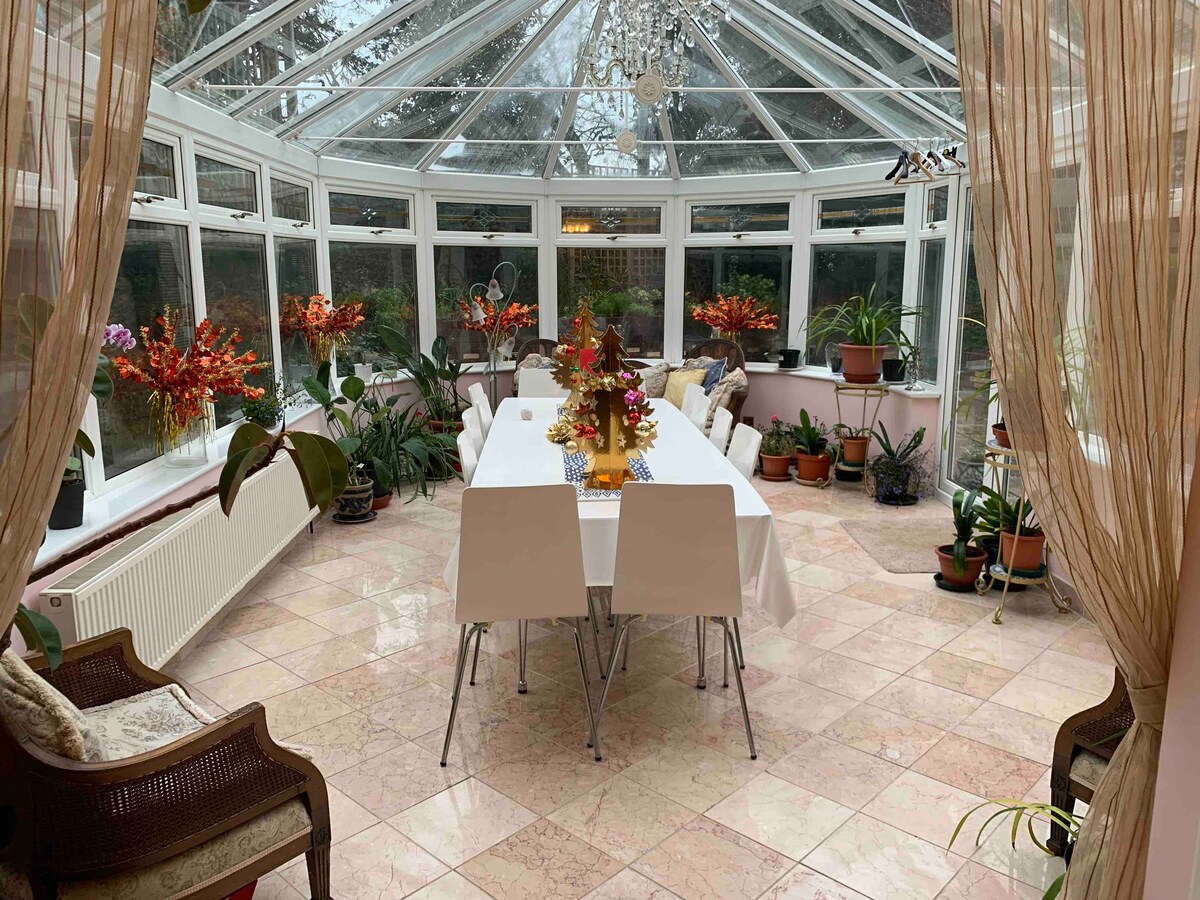 A bright conservatory filled with numerous potted plants along the windows and walls. A long dining table is set in the center, surrounded by white chairs. Natural light streams in through the glass roof and walls, creating a vibrant and uplifting space.
