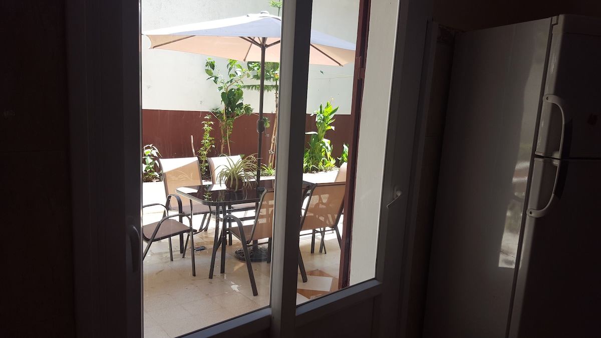 A private garden area is visible through the glass doors, featuring a round glass table surrounded by several chairs. A large umbrella provides shade, and potted plants offer greenery, creating a serene outdoor space attached to the kitchen.