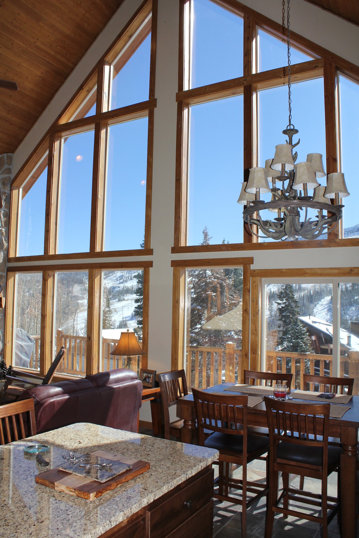 Large windows dominate the wall of this open space, allowing ample natural light and offering impressive views of the mountain landscape. A dining area with wooden chairs sits adjacent to a granite countertop, illuminated by a chandelier that complements the wood accents of the room.