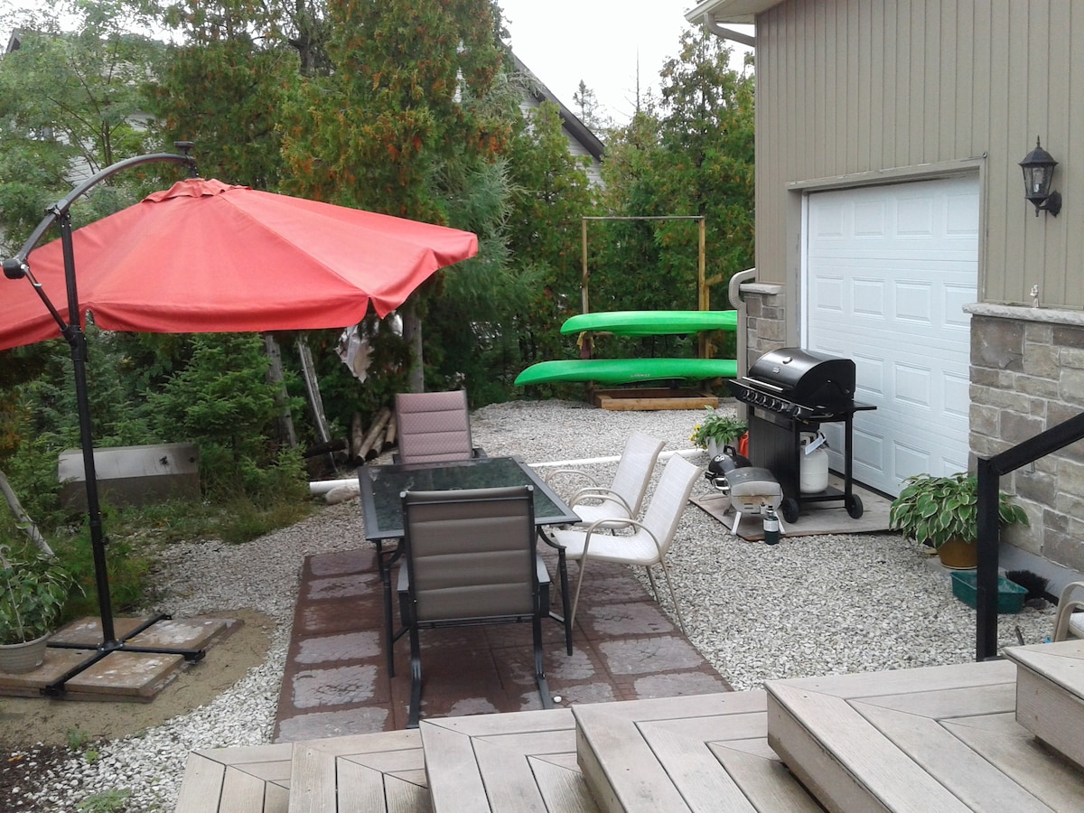 An outdoor patio area is depicted, featuring a glass-top dining table with four chairs. A large red umbrella provides shade. Nearby, a barbecue grill and two green kayaks are positioned against a backdrop of greenery, with a stone pathway leading towards the entrance.