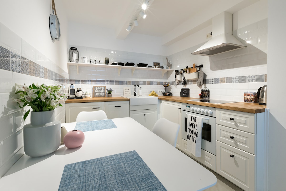 A modern kitchen presents white cabinetry with wooden countertops, enhancing a functional cooking space. Essential appliances, including a coffee machine and toaster, are visible. A dining table with a blue placemat is set for meals, while decorative plants add a touch of greenery.