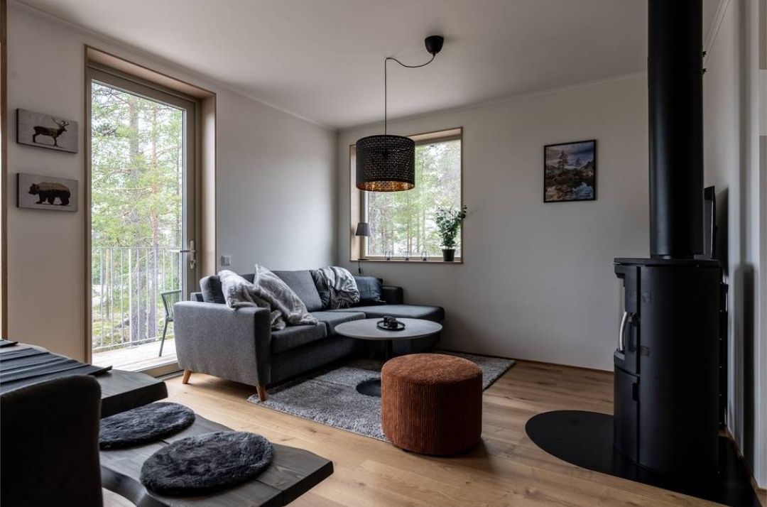 A modern living area features a comfortable gray sofa and a round coffee table, positioned on a textured rug. Large windows allow natural light to brighten the space, with a view of the outdoors. A stylish lamp hangs above, and a cozy accent ottoman complements the decor.