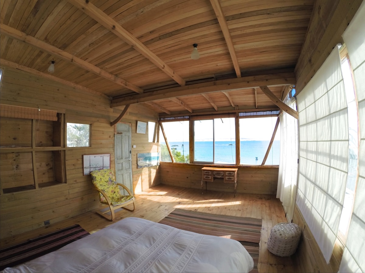 A bright and airy room features large windows that offer a panoramic view of the sea. The wood-paneled walls and ceiling provide a natural aesthetic. A spacious bed is positioned on a wooden floor, accompanied by a cozy chair and a small desk against the wall.