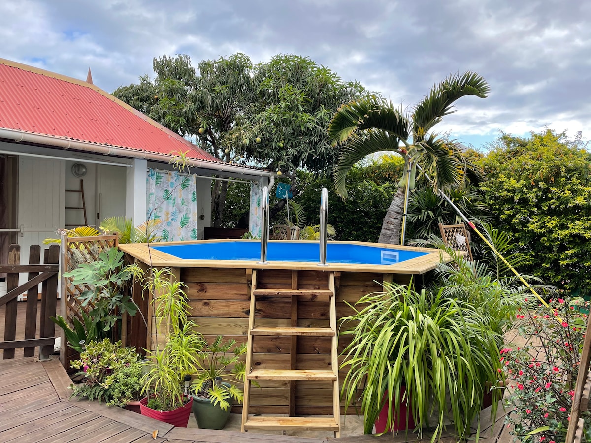 A wooden above-ground pool is surrounded by lush greenery and tropical plants. Steps lead up to the pool, which is framed by a spacious deck. A colorful wall in the background indicates the nearby bungalow. Cloudy skies provide a soft, diffused light.
