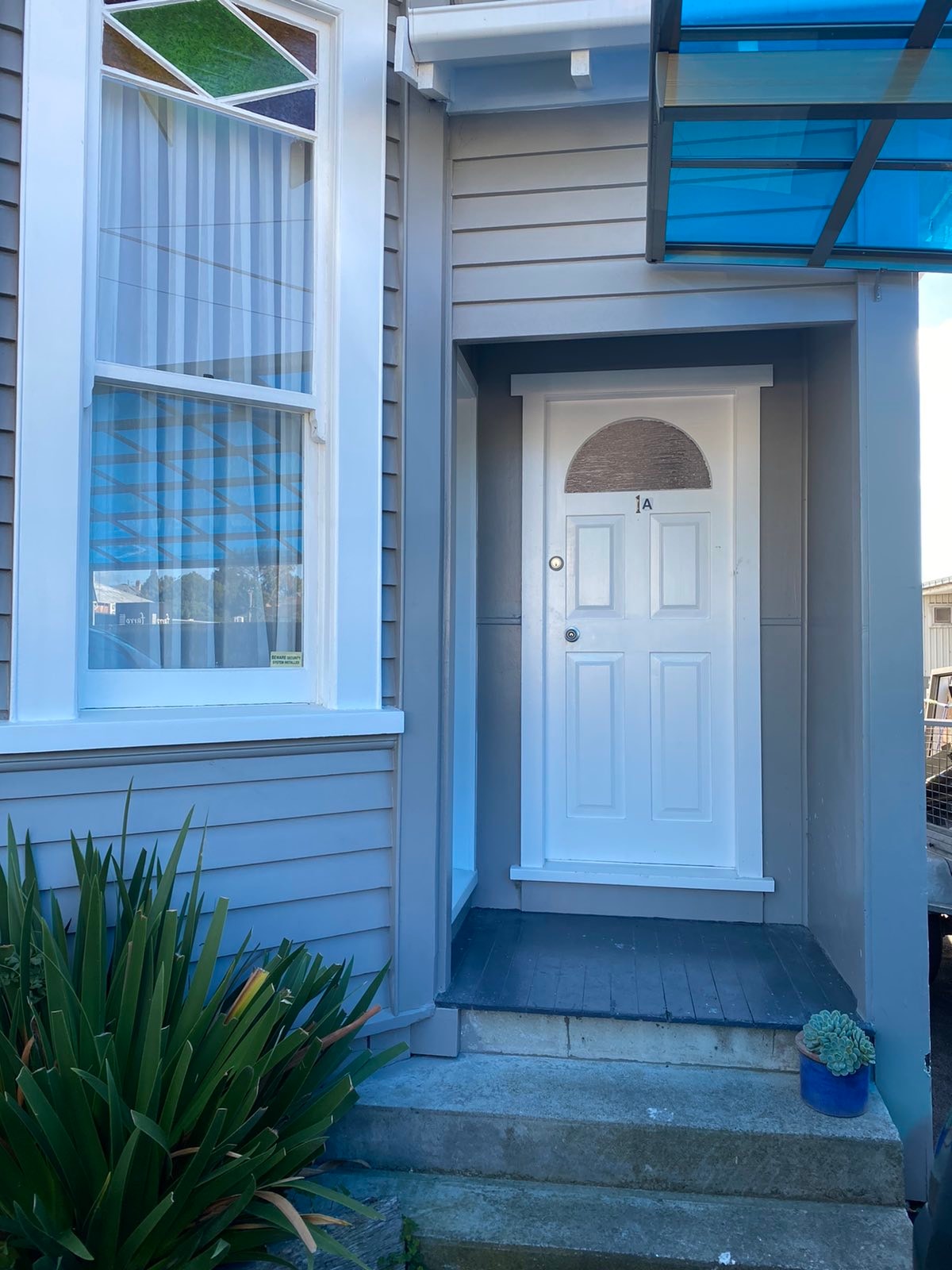 The entrance features a white front door with a circular window, surrounded by gray siding. A small set of steps leads to the door, and greenery is visible on both sides, providing a welcoming entryway.