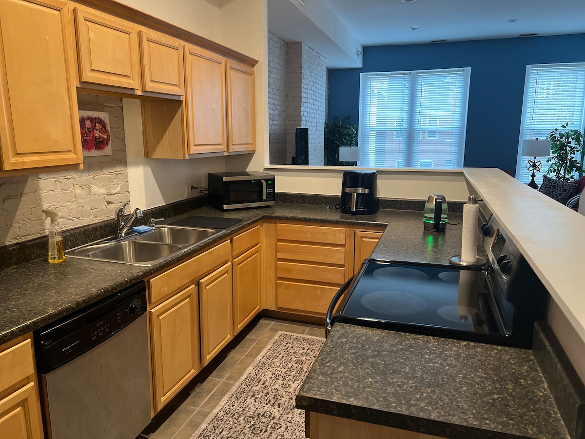 A fully equipped kitchen features light wood cabinetry and dark granite countertops. Stainless steel appliances include a dishwasher, oven, and microwave. A dual basin sink is positioned near a window, allowing natural light to brighten the space. Decorative accents add a touch of warmth.