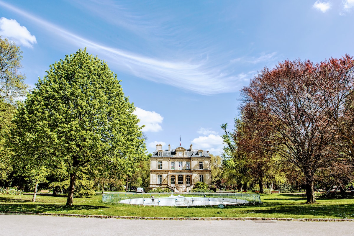 An elegant building is showcased surrounded by lush green trees, creating a serene outdoor environment. A fountain is featured in the foreground, complementing the well-maintained lawn and inviting setting under a clear blue sky.