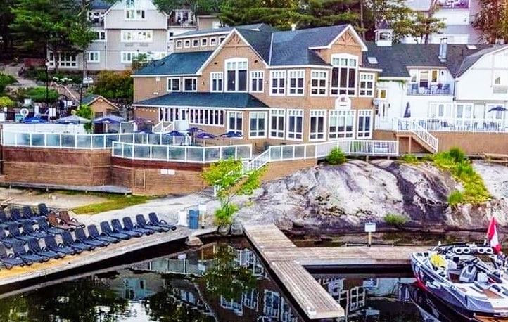 The exterior of a multi-story lodge is depicted, featuring a combination of large windows and a welcoming façade. A private dock extends into calm waters, with lounge chairs arranged in neat lines nearby. The surrounding landscape includes rocky formations and greenery.