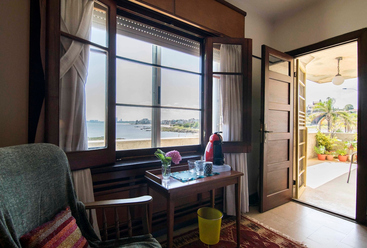 A cozy living area features a wooden table with a small vase of flowers and a kettle. Large windows offer expansive views of the beach and cityscape, while a doorway leads to a spacious terrace adorned with greenery.