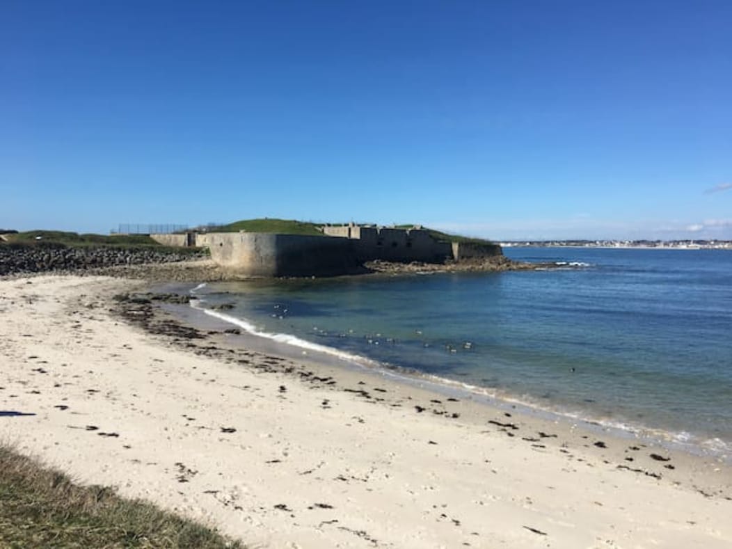 A sandy beach is bordered by gentle waves lapping at the shore, leading to a grassy area. In the background, the Fort de Porh Puns stands against a clear blue sky, surrounded by calm water. Seaweed is scattered along the tranquil beach.
