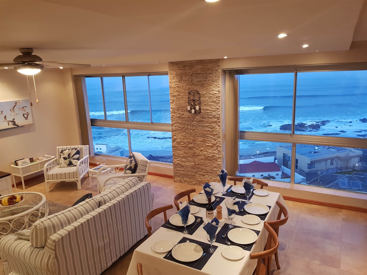 The spacious living area features a comfortable seating arrangement with a view of the ocean through large windows. A dining table is set for six, accompanied by vibrant blue tableware. Natural light enhances the stone accent wall, creating a serene atmosphere as waves crash below.