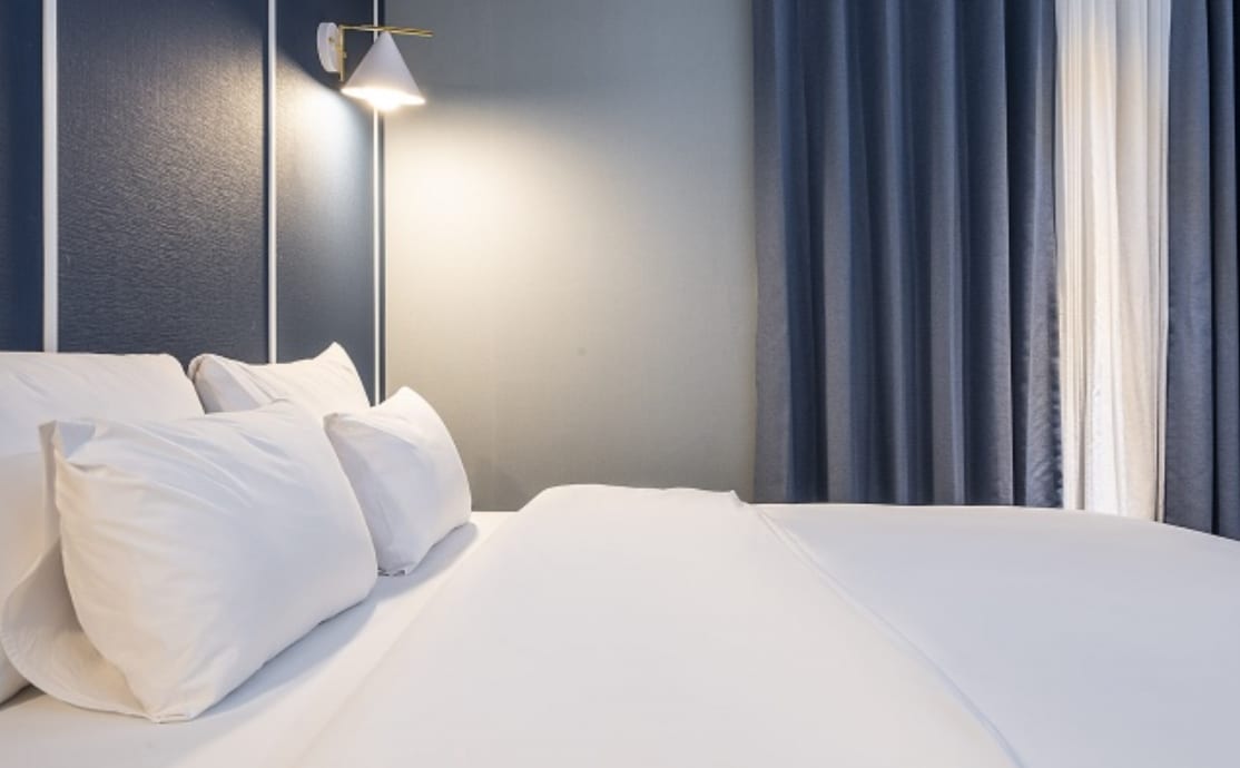 A cozy sleeping area features a spacious bed with neatly arranged pillows. A stylish wall-mounted lamp provides soft lighting, while floor-to-ceiling curtains frame a bright window, enhancing the inviting atmosphere.