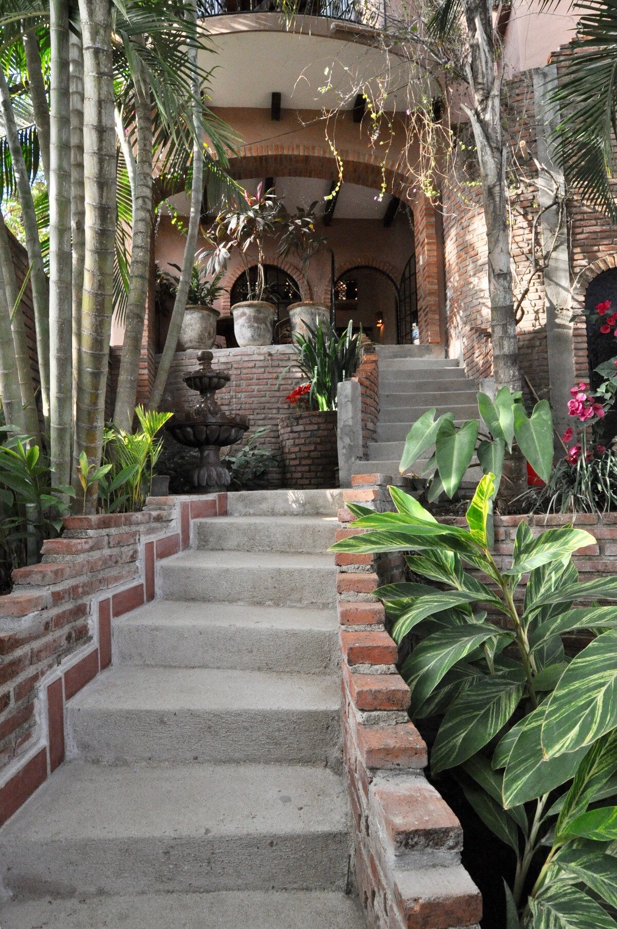 A set of stone steps leads up through a lush garden, flanked by ornamental plants and vibrant flowers. At the top, arched doorways reveal a terracotta and brick structure, complemented by decorative pots and a central fountain amidst the greenery.