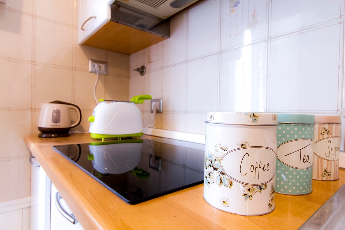 A well-equipped kitchen is presented, featuring a sleek black stovetop and light wooden countertops. A kettle and toaster in coordinating colors are visible, alongside decorative canisters labeled for coffee and tea. Subtle floral designs adorn the canisters, adding a touch of charm.