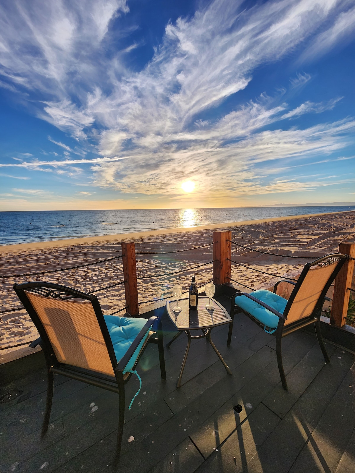 A cozy seating area is presented with two chairs, featuring blue cushions, facing the ocean. A small table is set between them, holding a bottle of wine. The backdrop highlights a vivid sunset over the water, casting warm hues across the sky.