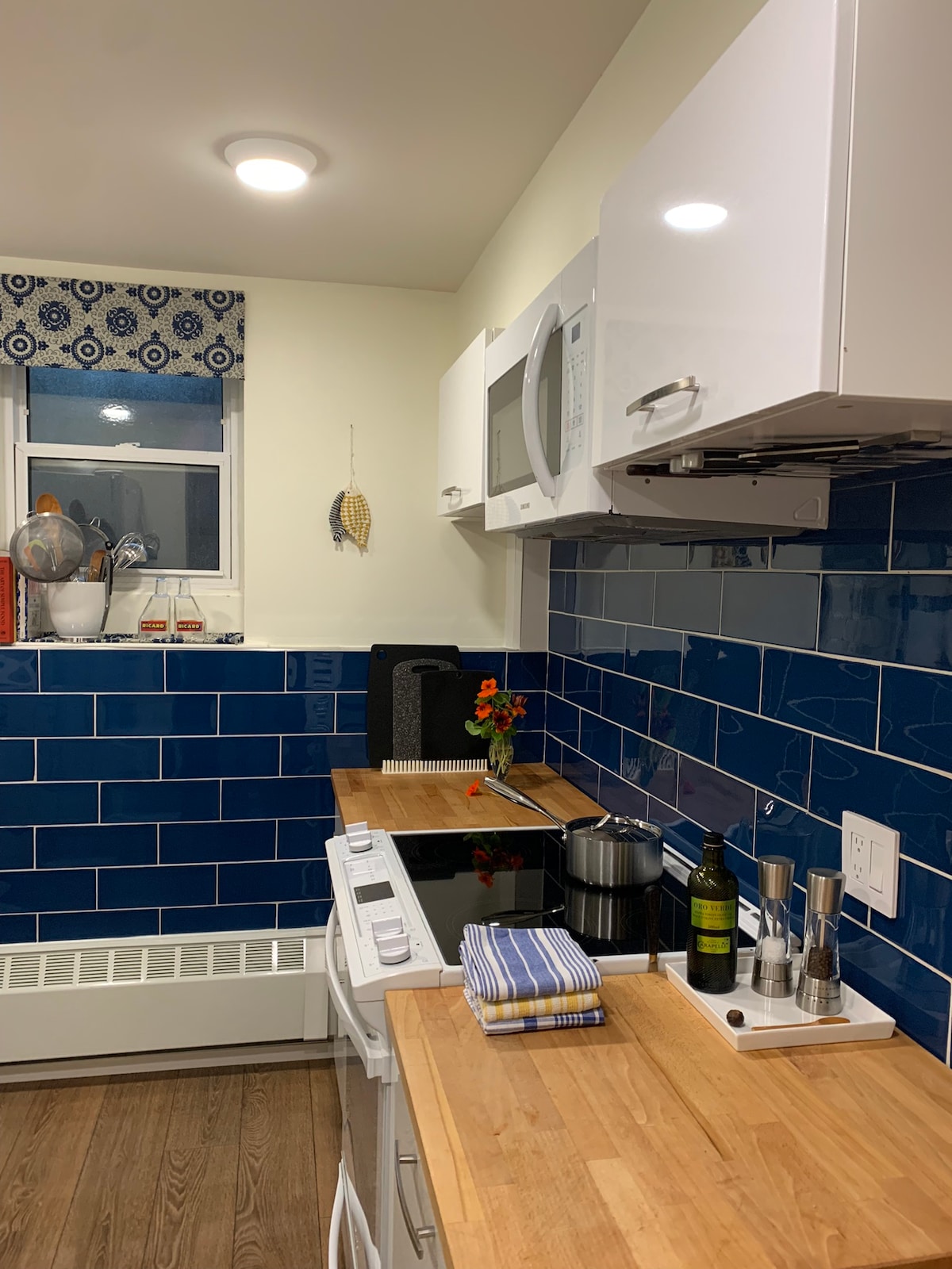 The kitchen features modern white cabinetry and deep blue tile backsplash, complemented by a wooden countertop. Cooking appliances, including an electric stove and microwave, are neatly arranged. A small vase with fresh flowers adds a pop of color, while the window provides natural light.