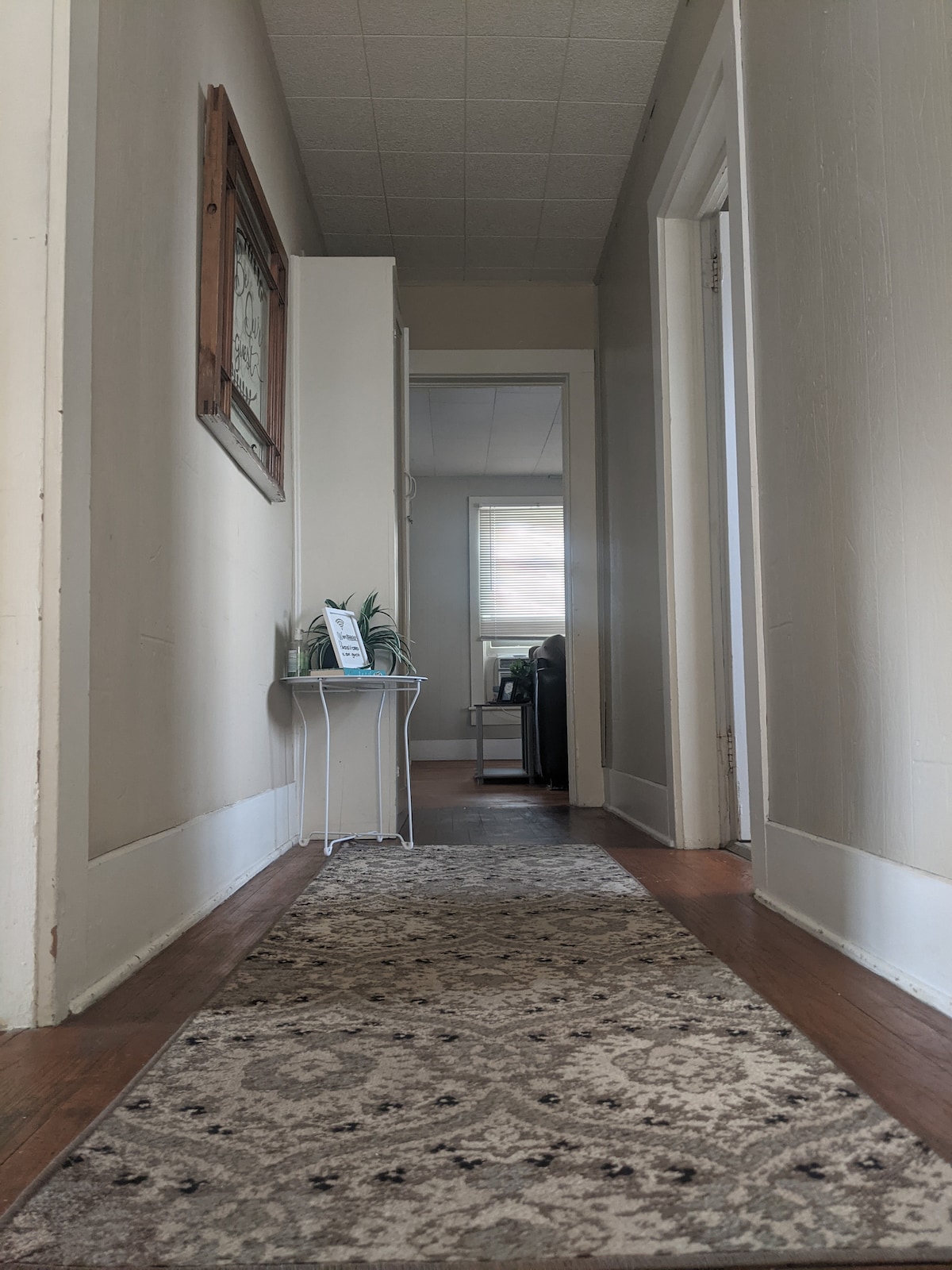 A long hallway is presented, featuring a patterned area rug and light-coloured walls. Framed artwork hangs on the left side. At the end of the hallway, a doorway leads into another room, with natural light coming through a window.