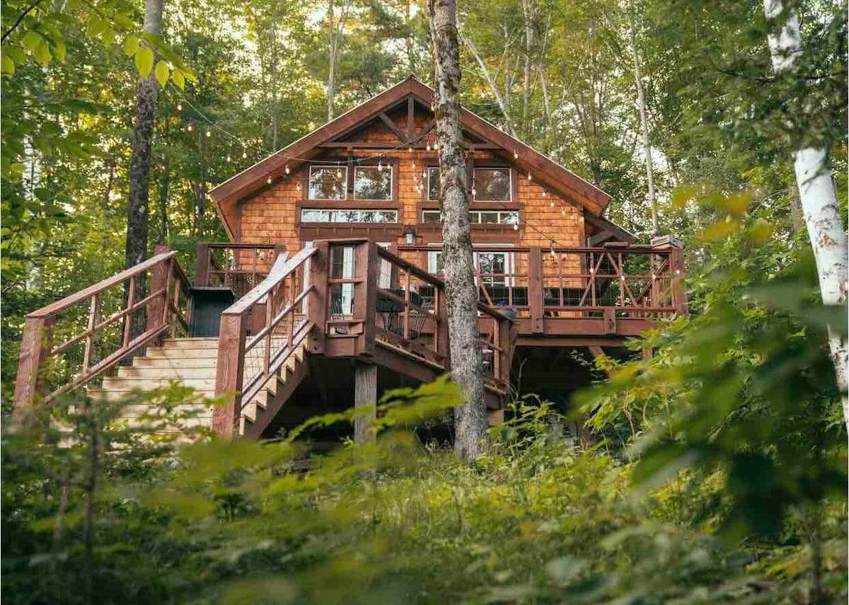 The treehouse, constructed of wood, is nestled among lush greenery. A two-story structure features large windows and a welcoming deck, surrounded by trees. Access is provided by a wooden staircase leading to the elevated platform, promoting a tranquil retreat setting in nature.